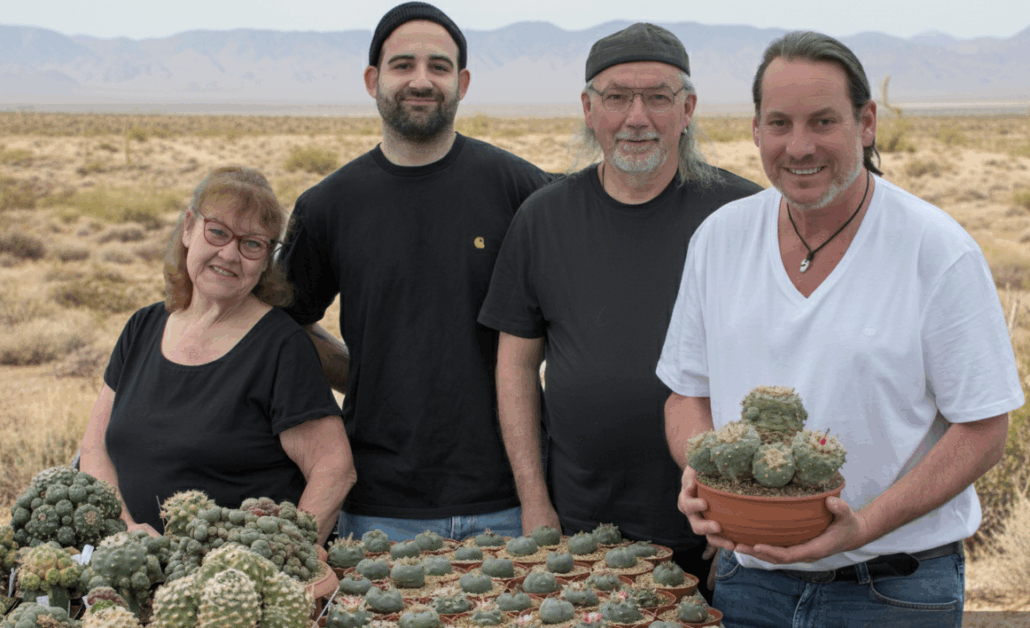 Das vierköpfige Team des Kakteenshop Lophophora präsentiert stolz eine große Sammlung verschiedener Lophophora-Kakteen in Töpfen vor einer Wüstenlandschaft.