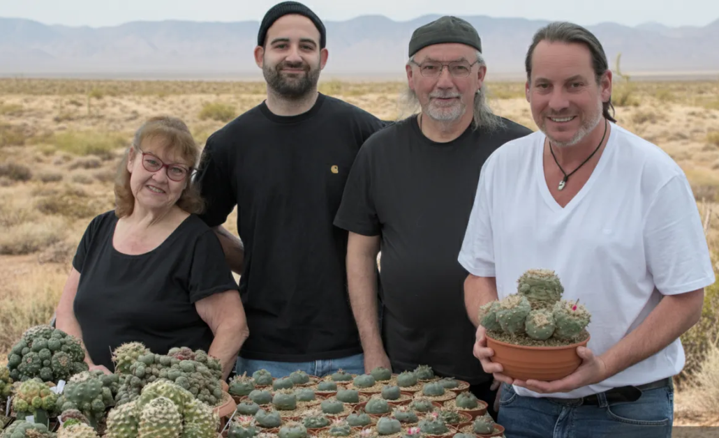 Das Team hinter Lophophora-williamsii.de – Ihre Kakteen-Experten Das vierköpfige Team des Kakteenshop Lophophora präsentiert stolz eine große Sammlung verschiedener Lophophora-Kakteen in Töpfen vor einer Wüstenlandschaft.